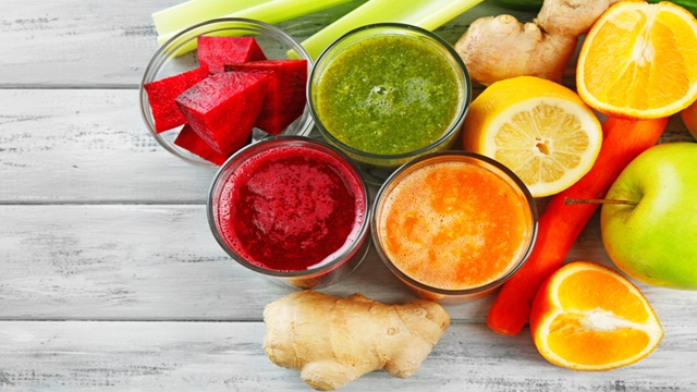 Four glasses of fresh vegetable and fruit juice in red, green, orange, and purple colors, surrounded by sliced beets, ginger root, celery stalks, carrots, apples, and citrus fruits on a rustic wooden surface